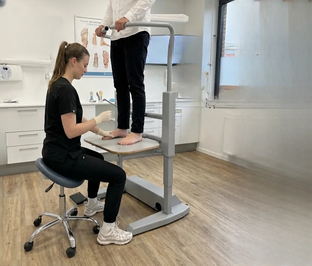 En fodplejespecialist sidder oprejst på en taburet med knæene under Orthostands hævede fodplade og undersøger den belastede fod på en klient, der står på liften og holder fast i gelænderet.