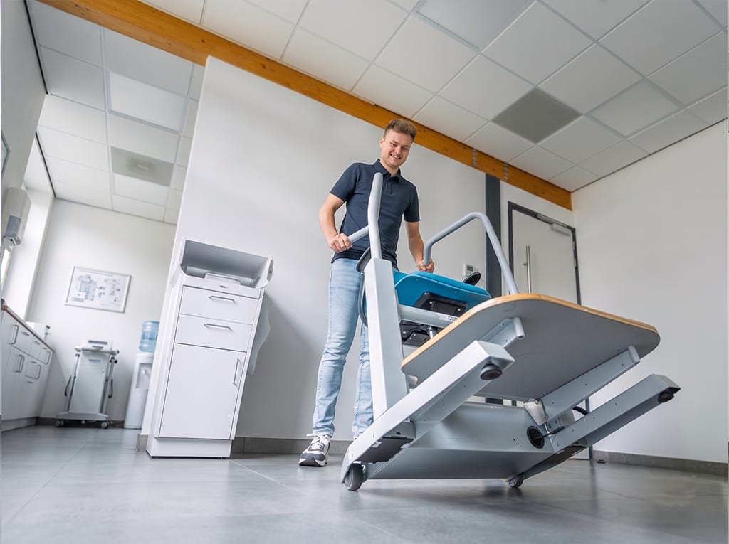 En behandler ruller Orthostand M30 hen over et behandlingsrum på hjulene, med platformen vippet i transportstilling